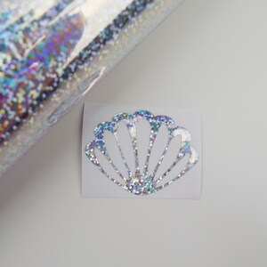 May include: A holographic glitter seashell sticker on a white background. The sticker is cut in the shape of a seashell with a shimmering, iridescent finish. A roll of holographic glitter material is visible in the background.