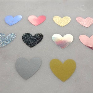 May include: Assortment of 10 heart-shaped embellishments in various colors and finishes, including glitter, iridescent, and solid colors. The hearts are approximately 2 inches in size.