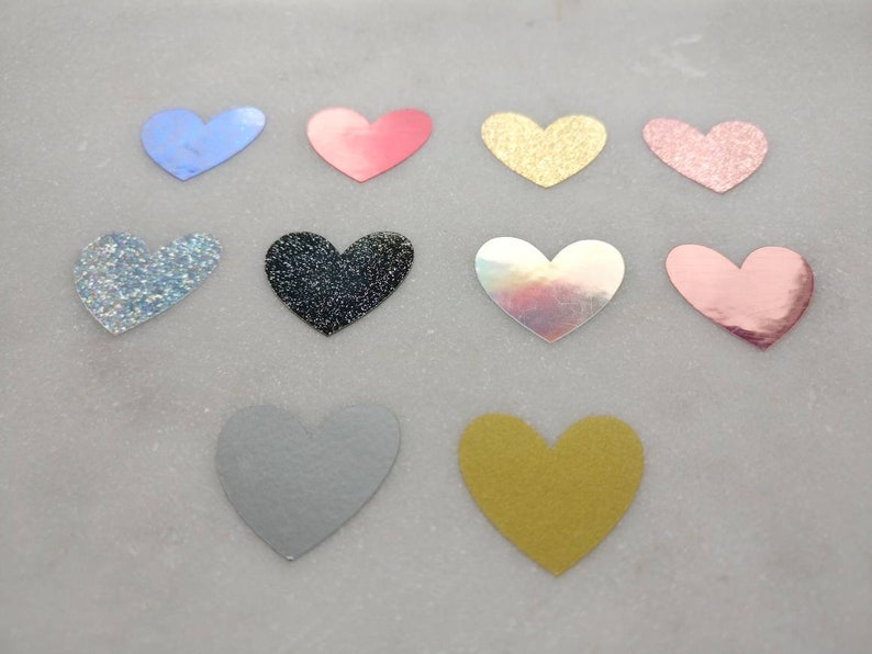 May include: Assortment of 10 heart-shaped embellishments in various colors and finishes, including glitter, iridescent, and solid colors. The hearts are approximately 2 inches in size.