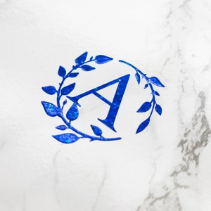 Può includere: Una lettera A blu glitterata circondata da una ghirlanda di foglie blu glitterate su uno sfondo di marmo bianco.