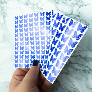 May include: Three sheets of blue glitter butterfly stickers. The stickers are arranged in a fan shape and are on a white background.