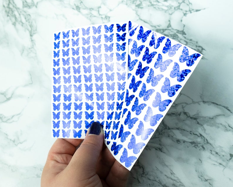 May include: Three sheets of blue glitter butterfly stickers. The stickers are arranged in a fan shape and are on a white background.