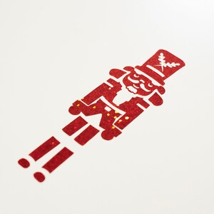 Könnte beinhalten: Roter Glitzer-Papp-Ausschnitt eines Nussknackers. Der Nussknacker blickt nach rechts und trägt einen hohen Hut mit einem Blatt-Design.