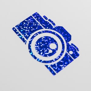 May include: A blue glitter camera icon cut out of white paper.