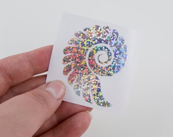 Pode incluir: Um adesivo branco com um design de concha de mar de glitter holográfico iridescente. A concha de mar tem a forma de uma espiral.