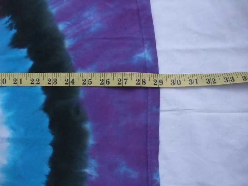 May include: A tie-dye textile with a color gradient of blue, black, and purple. A yellow measuring tape is laid across the fabric, showing a length of approximately 13 inches.