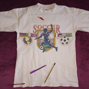 以下が含まれることがあります： サッカーをテーマにしたグラフィックが特徴の白いTシャツ。デザインには、サッカー選手のイラスト、サッカーボール、そして「WORLD CLASS INTERNATIONAL COMPETITION」というテキストの上に「SOCCER」という言葉が含まれています。このシャツは半袖です。
