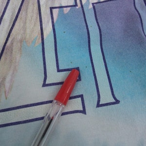 May include: Close-up of a light blue and white graphic tee with a red pen. The tee features a large, stylized letter "A" in dark blue, with a faded illustration in the background. The pen's red cap is visible.