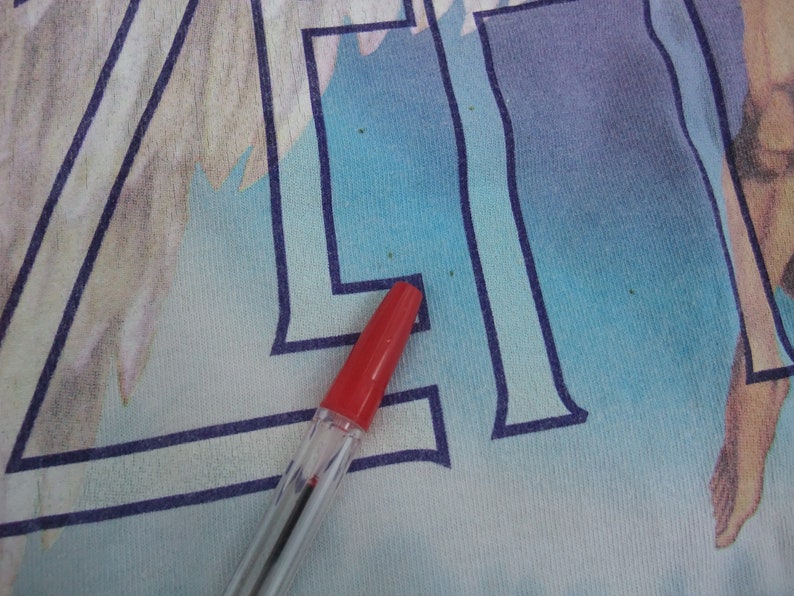 May include: Close-up of a light blue and white graphic tee with a red pen. The tee features a large, stylized letter "A" in dark blue, with a faded illustration in the background. The pen's red cap is visible.