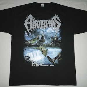 May include: Black t-shirt featuring the band name "Amorphis" in white, stylized lettering above a landscape graphic. The graphic depicts a waterfall and mountains. The text "Tales from + The Thousand Lakes" is below the graphic.