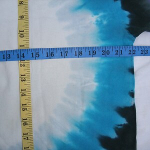 May include: White fabric with a tie-dye design in shades of blue, purple, and black. A yellow and blue measuring tape is laid across the fabric, showing measurements in inches.