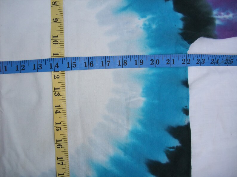May include: White fabric with a tie-dye design in shades of blue, purple, and black. A yellow and blue measuring tape is laid across the fabric, showing measurements in inches.
