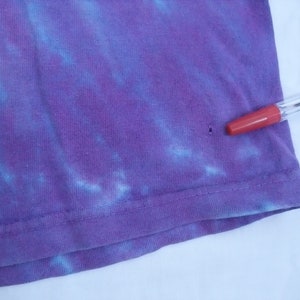 May include: Close-up of a purple tie-dye t-shirt with blue accents. The fabric is slightly wrinkled, and a red-tipped pen is resting on the shirt. The shirt has a small hole near the hem.