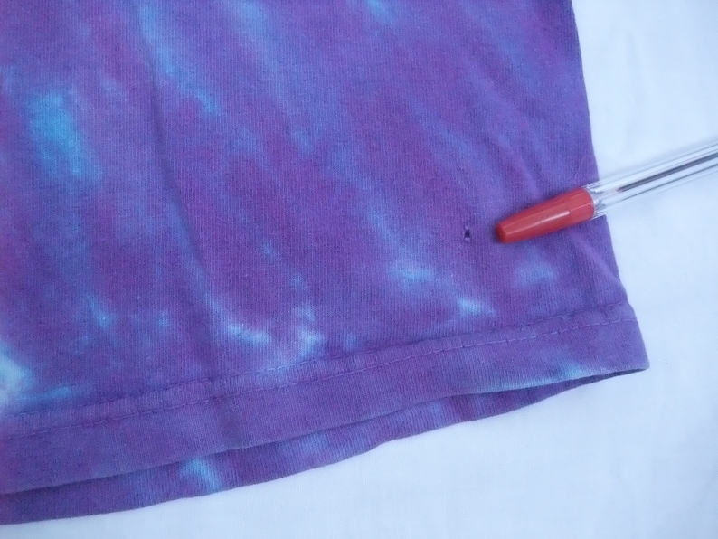 May include: Close-up of a purple tie-dye t-shirt with blue accents. The fabric is slightly wrinkled, and a red-tipped pen is resting on the shirt. The shirt has a small hole near the hem.
