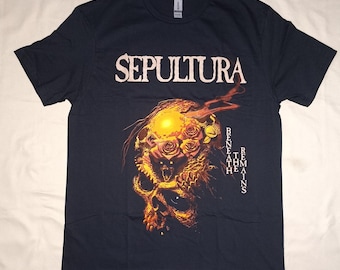May include: Navy blue t-shirt featuring the band name "SEPULTURA" in white block letters. Below the band name is a graphic of a skull with roses and flames. The text "BENEATH REMAINS" is printed vertically on the right side of the shirt.