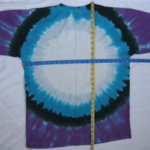 May include: A tie-dye t-shirt with a circular design in shades of purple, teal, black, and white. The shirt is laid flat, with a measuring tape showing a width of approximately 20 inches. The overall design is symmetrical.