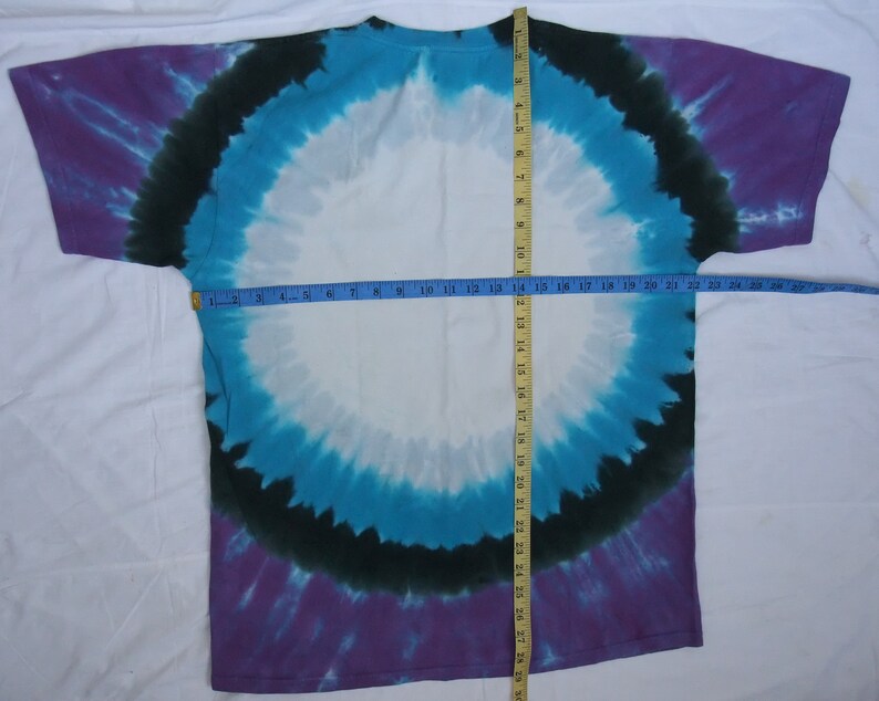 May include: A tie-dye t-shirt with a circular design in shades of purple, teal, black, and white. The shirt is laid flat, with a measuring tape showing a width of approximately 20 inches. The overall design is symmetrical.