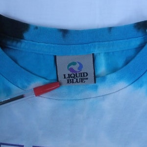 May include: Close-up of a blue tie-dye t-shirt collar with the "LIQUID BLUE" brand tag. The shirt has a light blue and white pattern. A red pen is in the foreground.