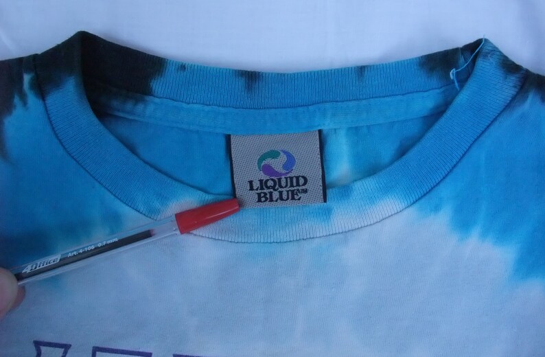 May include: Close-up of a blue tie-dye t-shirt collar with the "LIQUID BLUE" brand tag. The shirt has a light blue and white pattern. A red pen is in the foreground.