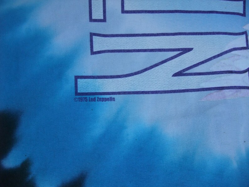May include: A blue tie-dye t-shirt with the Led Zeppelin logo in dark blue. The text "&copy;1975 Led Zeppelin" is printed at the bottom. The background has a cloudy, tie-dye effect.