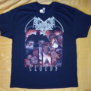 Può includere: T-shirt blu navy con un design gotico. Il design include un corvo, rose e un teschio, con la parola "CLOUDS" in scrittura bianca. Il nome della band "TIAMAT" è anch'esso visualizzato in lettere gotiche bianche.