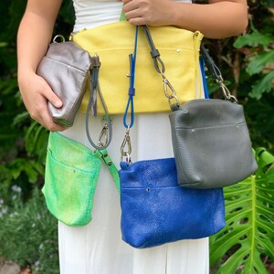 May include: A collection of colorful leather crossbody bags in various shades, including yellow, blue, green, and gray. Each bag features a textured finish and adjustable straps. The bags are displayed against a natural green background.
