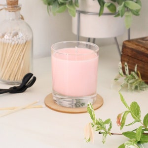 May include: A pink candle in a clear glass jar with a wooden coaster. The candle is lit and the wick is visible. The candle is on a white surface with a green plant in the background.