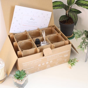 May include: An open cardboard box with a set of glass candle jars, wooden wicks, and a bottle of essential oil. The box is labeled "Time to Get Creative!" and has a list of social media icons. The box is sitting on a white surface with a small potted plant.