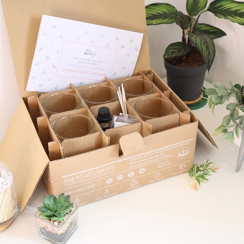May include: An open cardboard box with a set of glass candle jars, wooden wicks, and a bottle of essential oil. The box is labeled "Time to Get Creative!" and has a list of social media icons. The box is sitting on a white surface with a small potted plant.