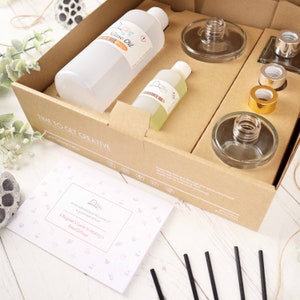 May include: A brown cardboard box with the words "TIME TO GET CREATIVE" printed on the front. The box contains a clear glass bottle with a white label that says "Base Oil", a smaller clear glass bottle with a yellow label that says "Fragrance Oil", a clear glass diffuser bottle, a set of black diffuser sticks, and a set of clear glass bottle stoppers. The box also includes a printed instruction booklet titled "A Beginner's Guide to Making a Reed Diffuser".