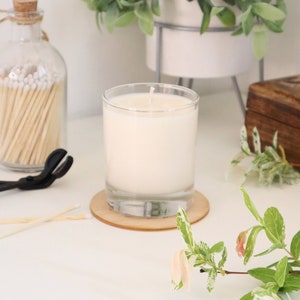 May include: A white candle in a clear glass jar with a wooden coaster. The candle is lit and the wick is visible. There are matches and a pair of tongs in the background.