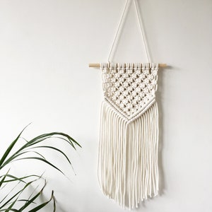 Op de afbeelding: Witte macramé wandhanger met een geometrisch patroon en franjes. De hanger is bevestigd aan een houten stok.