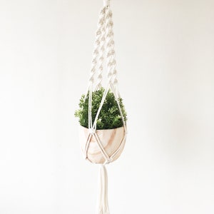 Op de afbeelding: Een witte macrame plantenhanger met een houten pot en een groene plant erin. De hanger hangt aan een haak aan het plafond.