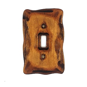 Puede incluir: Una cubierta de interruptor de luz de madera rústica con un acabado marrón desgastado. La cubierta tiene una forma ligeramente irregular y está diseñada para adaptarse a un interruptor de luz simple estándar.