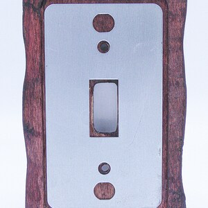 May include: A wooden light switch cover with a decorative, rustic design. The cover is dark brown with a light grey metal insert. The switch plate has a single toggle switch and two round openings.