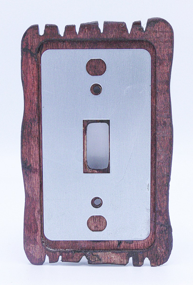 May include: A wooden light switch cover with a decorative, rustic design. The cover is dark brown with a light grey metal insert. The switch plate has a single toggle switch and two round openings.