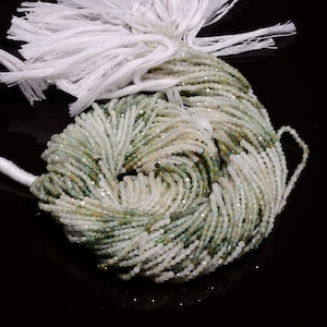 May include: A strand of white and green gemstone beads. The beads are small and round, and they are strung on a white thread. The beads are arranged in a loose coil.