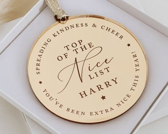 May include: A gold-colored, round Christmas ornament with the text "TOP OF THE NICE LIST HARRY" and the phrases "SPREADING KINDNESS & CHEER" and "YOU'VE BEEN EXTRA NICE THIS YEAR!" The ornament is in a white box with a gold ribbon.