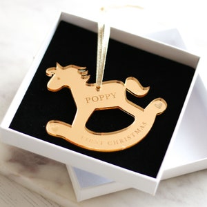 Puede incluir: Adorno de caballo de balanc&iacute;n de acr&iacute;lico de color dorado con el nombre "Poppy" y el texto "First Christmas" grabado en &eacute;l. El adorno est&aacute; en una caja blanca con un interior negro.