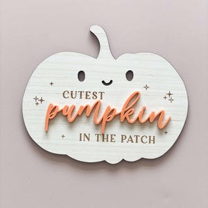 May include: A white wooden pumpkin with a cute face and the text "Cutest pumpkin in the patch" in brown lettering. The word "pumpkin" is in a raised, peach-coloured font.