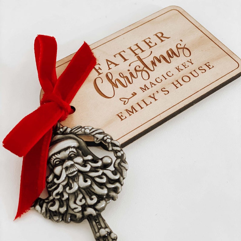 May include: A wooden tag with the text "Father Christmas Magic Key Emily's House" is tied to a silver key with a Santa Claus head. The key is tied with a red velvet ribbon.