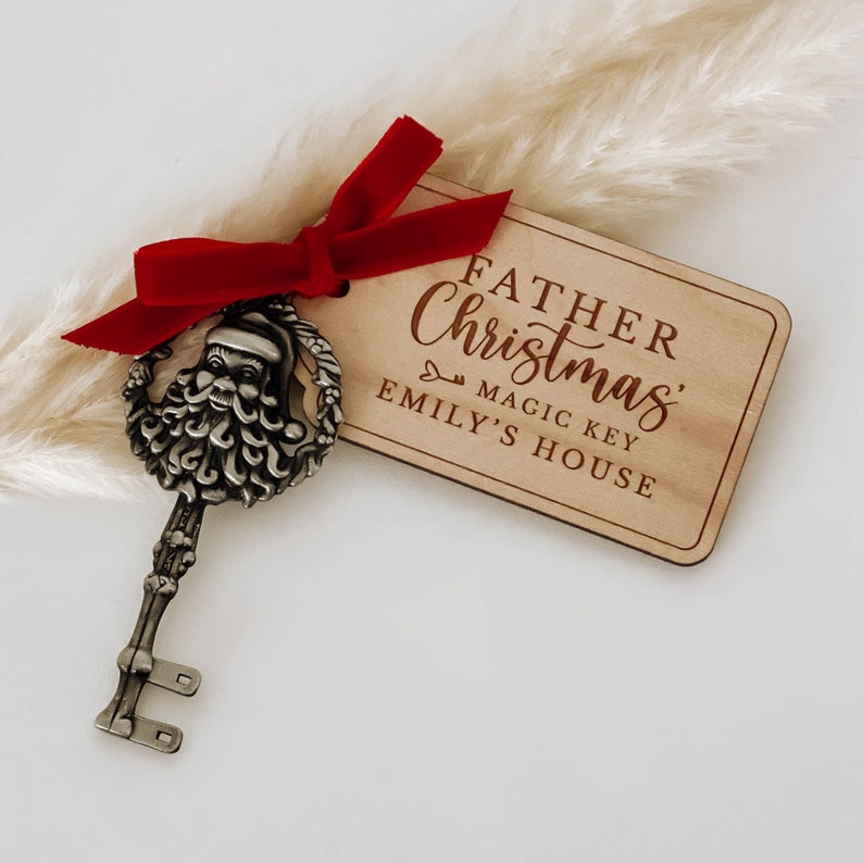 May include: A wooden tag with the text "Father Christmas Magic Key Emily's House" is tied to a silver key with a Santa Claus head. The key is tied with a red velvet ribbon.