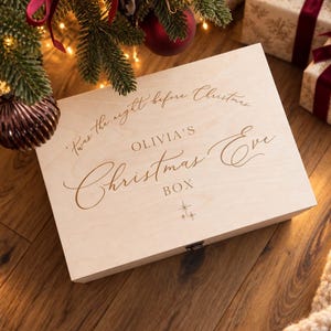 May include: A rectangular light-colored wooden box with a hinged lid and a metal latch. The lid features engraved text that reads 'Twas the night before Christmas', 'OLIVIA'S', 'Christmas Eve', and 'BOX', accompanied by a small star icon.
