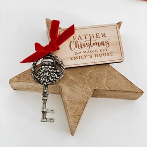 May include: A wooden star-shaped ornament with a silver key featuring a Santa Claus head. A red ribbon is tied around the key and the ornament. A wooden tag is attached to the ribbon with the text "Father Christmas Magic Key Emily's House".