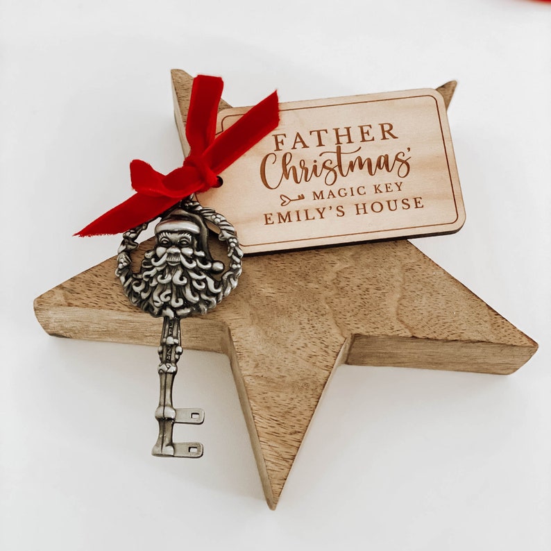 May include: A wooden star-shaped ornament with a silver key featuring a Santa Claus head. A red ribbon is tied around the key and the ornament. A wooden tag is attached to the ribbon with the text "Father Christmas Magic Key Emily's House".