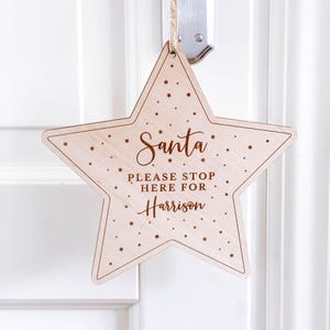 May include: A wooden star-shaped door hanger with the text "Santa Please Stop Here For Harrison" in a brown font. The star is decorated with small gold stars.
