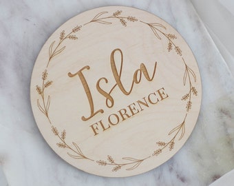 Może przedstawiać: Okrągła drewniana tabliczka w jasnym odcieniu z imieniem "Isla" w ozdobnej czcionce i nazwiskiem "Florence" poniżej, oba wygrawerowane. Wieniec kwiatowy otacza imiona, wszystko w jasnym odcieniu drewna.