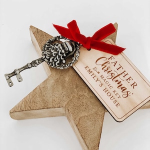 May include: A wooden star-shaped ornament with a silver key featuring a Santa Claus head. The key is attached to a red ribbon and a wooden tag that reads "Father Christmas Magic Key Emily's House".
