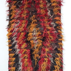 Peut inclure: Un tapis rectangulaire et touffu avec une base marron et un motif de touffes rouges, oranges, jaunes et noires. Le tapis est suspendu sur un fond blanc, maintenu par deux pinces à linge sur une baguette en bois.