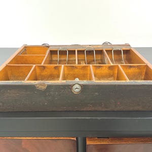 May include: A rectangular wooden cash drawer featuring a dark, weathered exterior finish and a central keyhole. The interior is divided into multiple compartments, with three metal spring-loaded bill clips positioned in the back row sections.